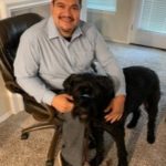 Photo of Esteban Rodriguez, sitting, with his large black dog, Goli, standing next to him.